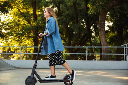 Photo of a cheerful beautiful happy young teenage girl outside in nature green park walking with scooter.の写真素材
