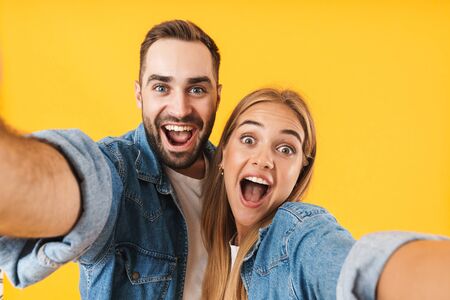 Image of amazed couple man and woman in denim clothes screaming at camera while taking selfie photo isolated over yellowの写真素材