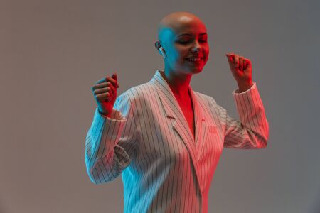 Portrait of an attractive bald woman wearing jacket standing isolated over gray background under red light, listening to music with wireless earphonesの写真素材