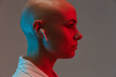 Side view portrait of an attractive bald woman wearing jacket standing isolated over gray background under red light, listening to music with wireless earphonesの写真素材