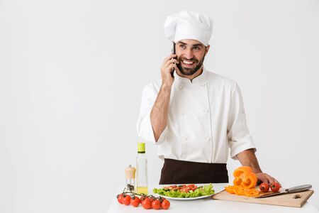 Image of happy chef man in uniform smiling and talking on smartphone while cooking vegetable salad...