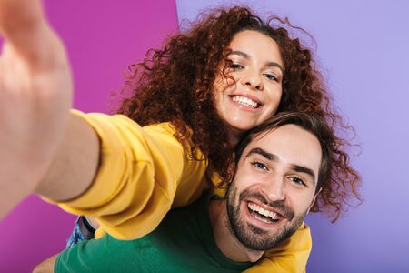 Portrait of joyful smiling couple man and woman in colorful clothing taking selfie photo while doing piggyback ride isolated over purple backgroundの写真素材