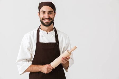 Image of professional chief man in cook uniform smiling and holding kitchen wooden rolling pin isolated over white wallの写真素材