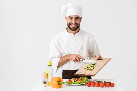 Image of cheerful cook man in uniform smiling and cutting vegetable salad on wooden board isolated...