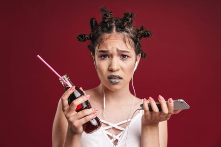 Image of puzzled punk girl wearing earphones expressing wonder while holding soda bottle and smartphone isolated over red backgroundの写真素材