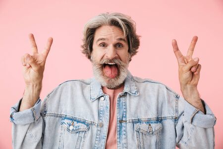 Portrait of a cheerful middle aged man wearing casual outfit standing isolated over pink background, showing peace gestureの写真素材