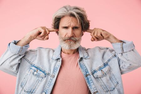 Portrait of an annoyed middle aged man wearing casual outfit standing isolated over pink background, covering earsの写真素材