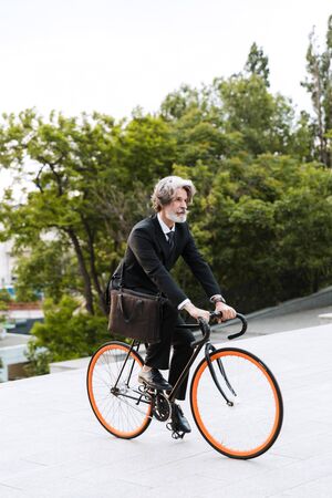 Handsome confident bearded man wearing suit, carrying briefcase outdoors riding on a bicycleの写真素材