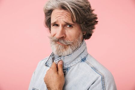 Portrait of mustached old man with gray beard wearing denim jacket looking at camera isolated over pink backgroundの写真素材