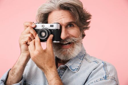 Portrait of satisfied old man with gray beard wearing denim jacket smiling and taking photo at retro...