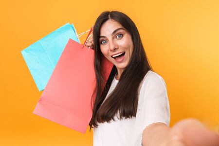 Portrait of excited shopaholic woman wearing dress smiling and taking selfie photo while holding colorful shopping bags isolated over yellow backgroundの写真素材