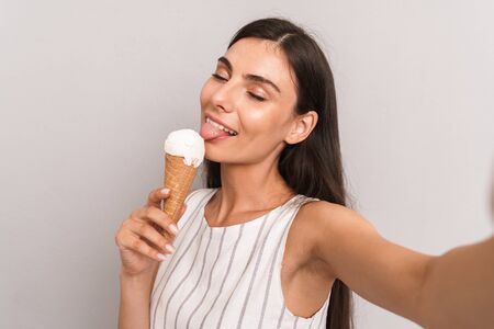 Image of young brunette woman wearing dress smiling and taking selfie photo while eating ice cream...