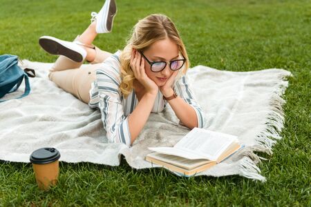 Beautiful young blonde girl relaxing on a lawn at the park, reading a book while laying on a blanketの写真素材