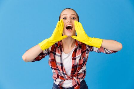 Portrait of lovely housewife wearing yellow rubber gloves screaming at camera while doing housework and cleaning room isolated over blue backgroundの写真素材