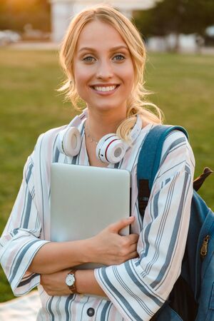 Happy young blonde girl carrying backpack and laptop computer standing at the park