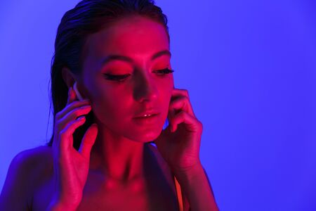 Image of beautiful sensual woman wearing bodysuit underwear listening to music with earpods isolated over violet neon backgroundの写真素材