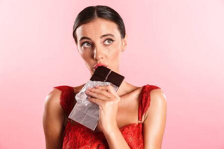 Closeup image of cute thinking woman wearing red sexual lace lingerie eating chocolate bar closed isolated over pink backgroundの写真素材