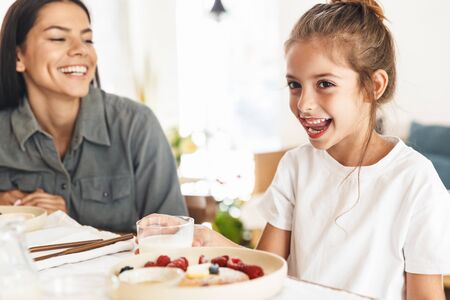 Image of amazing family mother and little daughter having breakfast at home in morning while sitting at table in bright kitchenの写真素材
