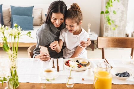 Image of caucasian family mother and little daughter hugging and using cellphone together while having breakfast at home in morningの写真素材
