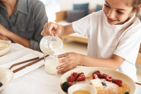 Image of beautiful family mother and little daughter having breakfast at home in morning while sitting at table in bright kitchenの写真素材