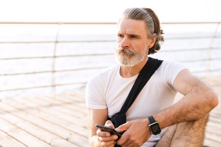 Image of serious elderly man in sportswear typing on cellphone while sitting near seaside in morningの写真素材