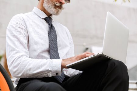 Cropped photo of handsome gray-haired businessman in formal clothes typing on laptop while sitting outdoorsの写真素材