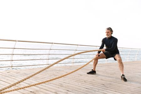 Image of strong old man in tracksuit doing exercise with battle ropes while working out near seaside...