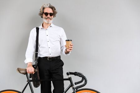 Photo of joyful mature businessman in sunglasses drinking coffee takeaway while standing with...