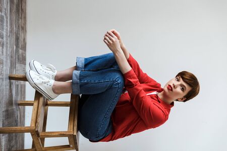 Image of brunette young woman looking at camera while sitting on wooden stand isolated over gray...
