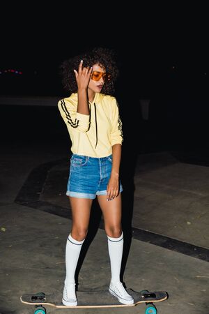 Image of a young amazing african woman in sunglasses at night walking by street on skateboard.