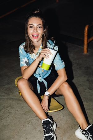 Image of positive brunette girl in streetwear drinking soda and sitting on skateboard at night outdoorsの写真素材
