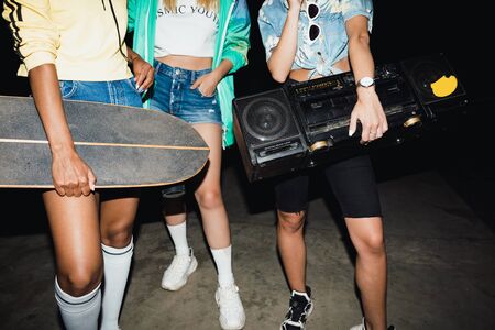 Cropped image of stylish girls walking with skateboards and boombox player at night outdoorsの写真素材