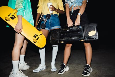 Cropped image of young girls walking with skateboards and boombox player at night outdoors