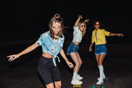 Image of fashion multinational girls in streetwear smiling and riding skateboards at night outdoors