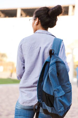 Back view of an attractive young african woman student carrying backpack walking outdoors, listening to music with wireless earphonesの写真素材
