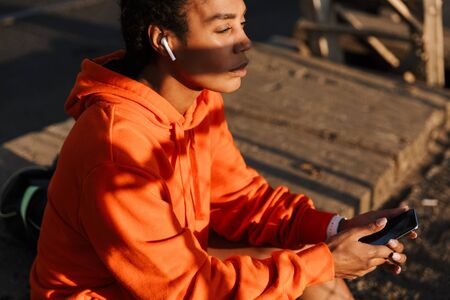 Photo of african american concentrated woman in sportswear using earpods and cellphone while sitting on old bridgeの写真素材