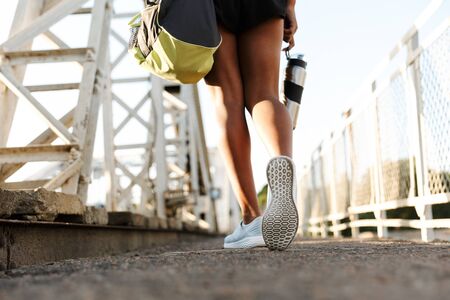 Cropped photo of african america woman in sportswear holding water bottle while walking with bag on old bridgeの写真素材