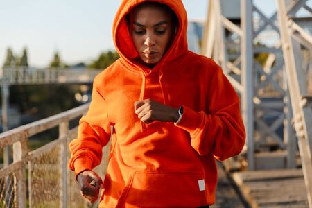 Photo of african american serious woman in hoodie running while working out on old bridgeの写真素材