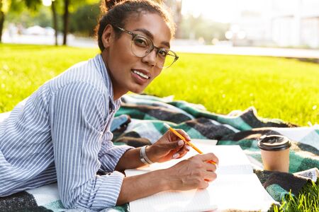Image of happy african american woman studying with exercise books while lying on blanket in parkの写真素材