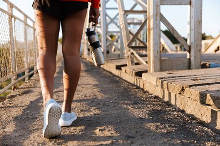 Cropped photo of african american woman in sportswear holding water bottle while walking on old bridgeの写真素材