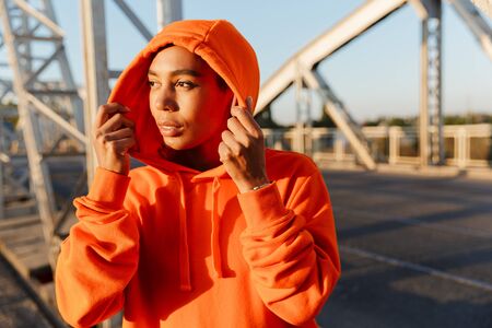 Photo of african american pleased woman in hoodie using earpods while working out on old bridge