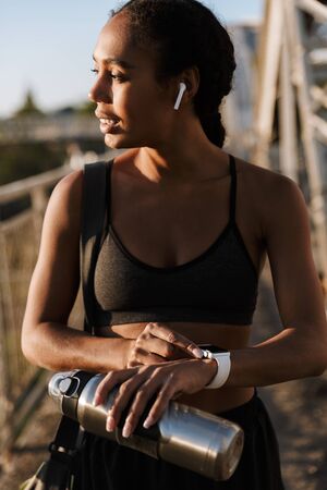 Photo of african american happy woman in sportswear using earpods and smartwatch while walking on old bridgeの写真素材