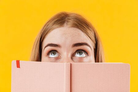 Image of a young girl isolated over yellow wall background covering face with notebook looking aside.の写真素材