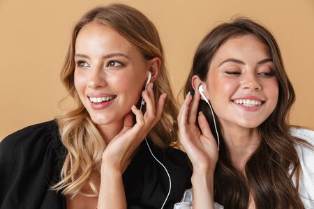 Portrait closeup of two cheerful women listening music with earphones isolated over beige backgroundの写真素材
