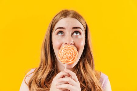 Image of a cheery surprised happy young cute teenage girl posing isolated over yellow wall background holding sweet candy lollipop covering mouth looking aside.の写真素材