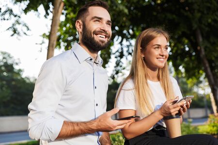Photo of a young cheerful happy man and woman businesspeople outside in nature park using mobile...
