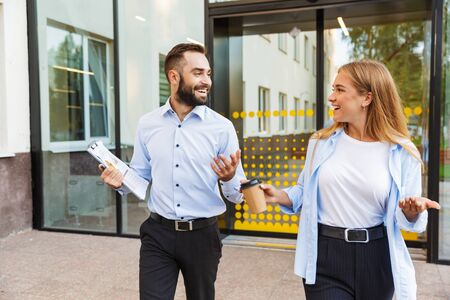 Image of a young happy man and woman businesspeople outside at the street near business center holding clipboard and cup of coffee talking with each other.の写真素材