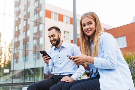 Image of a young happy colleagues man and woman businesspeople sit outside at the street using...