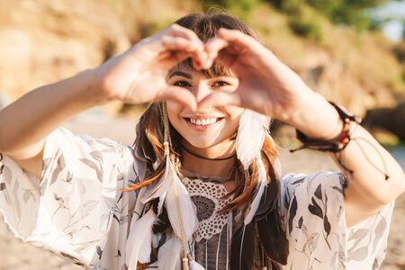 Image of lovely hippy girl in feather headband showing heart shape while walking by seaside in morningの写真素材