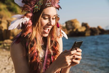 Image of excited hippy girl in indian chief feather headdress holding smartphone while walking by...